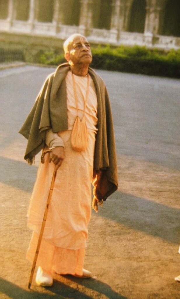 AC Bhaktivedanta Swami Srila Prabhupada standing on an open pathway in saffron robes and a grey woolen shawl, holding a wooden walking stick, wearing tulasi beads and a japa bag, gazing peacefully upward toward the sky. Lush greenery and a white ornamental structure visible in the background. Warm golden light suggests early morning or evening — the sacred hours of bhajan.